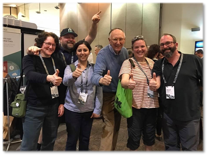 Photograph of blog post contributor Harkeerat Kang posing with work colleagues at an Esri conference, all showing thumbs up along with Esri co-founder Jack Dangermond